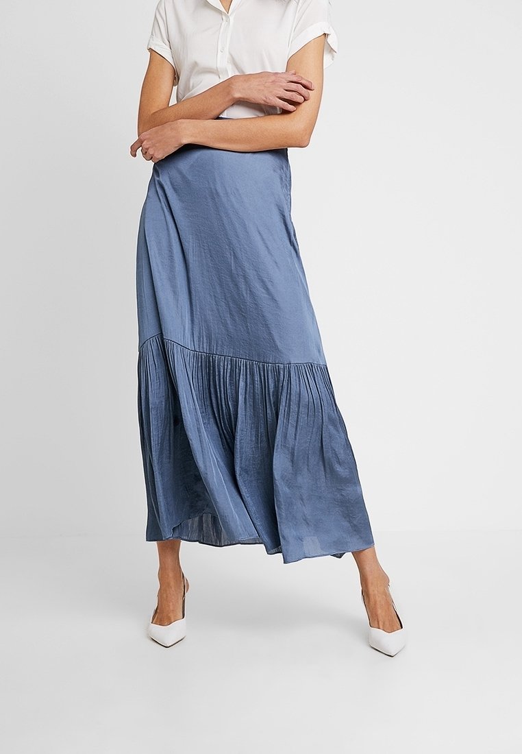 Kvinna i vit kortärmad blus och en flödande blå midikjol med vita spetsiga klackar, stående med armarna i kors.