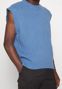 Gilet sans manches en tricot bleu clair porté avec un pantalon noir, les mains du modèle dans les poches, sur un fond blanc uni.