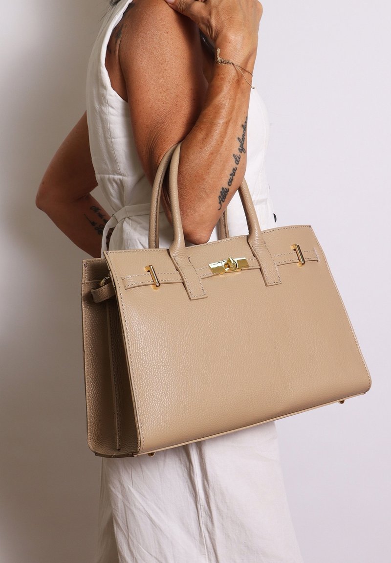 Sac fourre-tout en cuir beige avec un design structuré, deux poignées, des détails en métal doré et une finition texturée. Silhouette compacte et moderne.