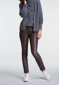 Bruine leren broek met een slim fit, gecombineerd met een grijze gebreide trui en witte sneakers. De trui heeft een hoge kraag en decoratieve elementen.