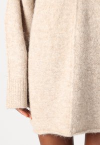 Beige neulemekko, jossa on pehmeä ja pörröinen rakenne, pitkät hihat ja hieman kaareva helma. Ribbattu yksityiskohta hihansuissa.