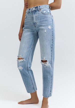 Kobieta ubrana w jasnobłękitne jeansy z wysokim stanem, z postrzępionymi dziurami na kolanach, stoi boso na jednolitym tle.