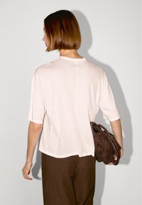 Korte mouwen, lichtroze T-shirt met een relaxte pasvorm. Inclusief een centrale achternaad. Gedragen met bruine broek en een gevlochten bruine tas.