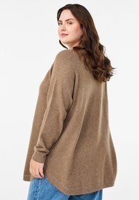 Brūns adīts džemperis ar brīvu piegriezumu, garām piedurknēm un ribotām maliņām. Kombinēts ar gaiši ziliem džinsiem, kuriem ir noapaļots apakšmalas dizains.