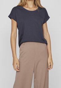 Žena v námořnické modré tričku s krátkým rukávem a ohrnutými manžety, zasunutém do béžových kalhot s vysokým pasem a širokými nohavicemi, na jednobarevném pozadí.