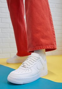 White leather sneakers with perforated toe, textured sole, and laces, paired with red wide-legged pants. Background features blue and yellow surfaces.