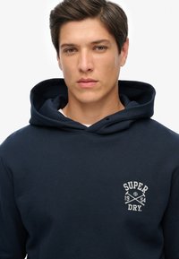 Felpa blu navy con tasca anteriore, cappuccio con lacci e un logo bianco che presenta "SUPER DRY" e asce incrociate sul lato sinistro del petto.