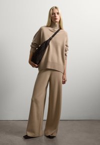 Donna che indossa un maglione beige a collo alto e pantaloni a gamba larga, che tiene una borsa a tracolla nera, in piedi contro una parete bianca liscia.