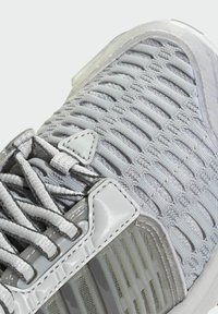 Gros plan d'une chaussure de sport grise avec une tige en mesh texturé, un système de lacets et des détails de support en plastique translucide près de la semelle.