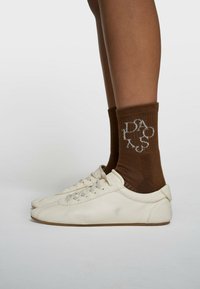 Baskets blanches texturées avec lacets, associées à des chaussettes marron côtelées ornées d'accents de logo blancs, présentées sur un fond neutre.