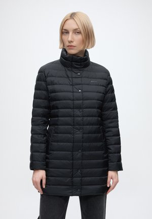 Frau mit kurzen blonden Haaren, die einen schwarzen gesteppte Wintermantel mit Knöpfen und dunkle Hose vor einem schlichten weißen Hintergrund trägt.