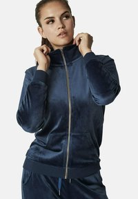 Veste zippée en velours bleu marine avec col haut, poignets côtelés et poches avant. Texture lisse, design épuré et détail de fermeture éclair métallique.