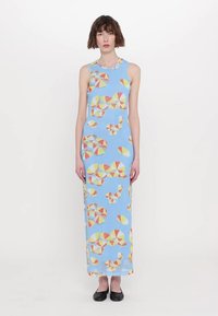 Robe longue sans manches avec un fond bleu présentant un motif géométrique en jaune, orange et rouge. Le modèle porte des chaussures noires.