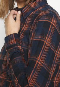 Camicia in flanella a quadri blu navy e arancione, con colletto a polo, maniche lunghe e tessuto strutturato. Linee orizzontali e verticali notevoli.