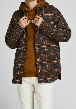 Camicia a scacchi marrone con accenti blu e arancioni, design con bottoni, indossata sopra un hoodie marrone. Pantaloni bianchi completano l'outfit.