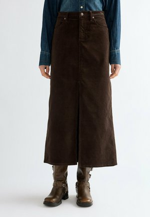 Persona que lleva una falda larga marrón oscuro con abertura frontal, camisa vaquera y botas de cuero marrón hasta media pantorrilla con correas y hebillas.