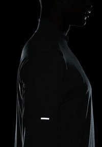 Camisa deportiva de manga larga en gris oscuro con una textura suave; cuenta con un acento reflectante blanco en la parte superior del brazo.