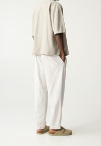 Chemise beige à manches courtes, coupe décontractée, associée à un pantalon large à rayures blanches. Chaussures : mocassins marron avec une boucle.