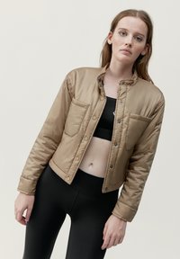 Tan quiltad jacka med tryckknappar, två bröstfickor och hög krage. Bärs över en svart topp och leggingar, med en slät textur.