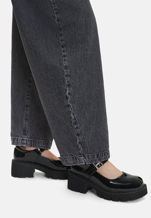 Czarne lakierowane buty typu Mary Jane z masywną podeszwą i regulowanym paskiem. Połączone z szerokimi szarymi spodniami z denimu.