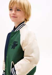 Blondes Kind mit Sommersprossen, das eine grüne, marineblaue und cremefarbene College-Jacke mit gestreiftem Kragen und Bündchen trägt, die Hände in den Taschen, schaut leicht nach oben.