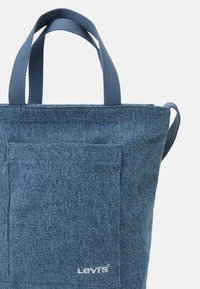 Denim tote bag in blue with two textile handles, a front pocket, and a white logo "LEVI'S" on the lower right. Textured surface.