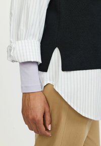 Zoom su una mano accanto a dei pantaloni beige, una camicia a righe bianca e grigia con maniche arrotolate, e un maglione senza maniche nero con uno spacco laterale.