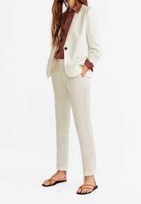 Licht beige linnen pak met een blazer met één knoop en een op maat gemaakte broek. Aangevuld met een donkerbruine overhemd met knopen en platte sandalen.