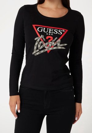 Vrouw die een nauwsluitend zwart shirt met lange mouwen draagt, met een rood driehoekig en versierd "Guess Icon" logo op de voorkant.