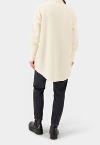 Persona che indossa una giacca di pile color crema, pantaloni neri e stivali con piattaforma neri, in piedi con la schiena rivolta verso l'osservatore su uno sfondo bianco.