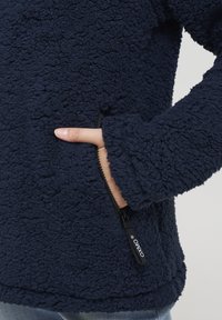 Giacca in pile blu navy con superficie texture, tasca con zip e polsini elasticizzati. Presenta un'etichetta con logo vicino alla zip.