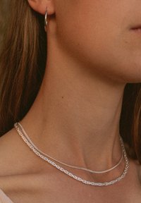 Silberne Halsketten, die übereinander gelegt um den Hals getragen werden, mit einem strukturierten Kettendesign und einer glatten, dünnen Kette, wobei ein kleiner Creolenohrring sichtbar ist.