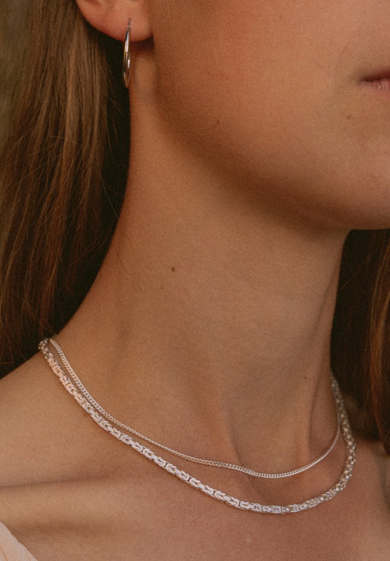 Silberne Halsketten, die übereinander gelegt um den Hals getragen werden, mit einem strukturierten Kettendesign und einer glatten, dünnen Kette, wobei ein kleiner Creolenohrring sichtbar ist.
