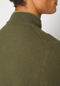 Lähikuva henkilöstä, joka on pukeutunut oliivinvihreään ribbineuleeseen, jossa on korkea kaulus. Näkymä takaa ja sivulta.