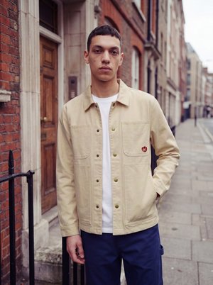 Jeune homme debout sur un trottoir en ville portant une veste beige par-dessus une chemise blanche et un pantalon bleu, une main dans la poche.