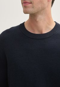 Maglione lavorato a maglia blu navy con scollatura a girocollo a coste e un tessuto testurizzato. La vista ravvicinata mette in evidenza i dettagli della scollatura e delle cuciture.