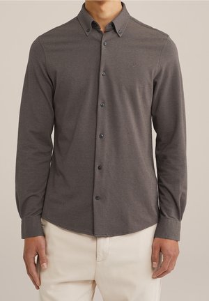 Uomo che indossa una camicia grigio scuro a maniche lunghe abbottonata con collo e pantaloni beige chiaro, in piedi contro uno sfondo semplice.