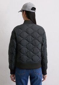 Czarna pikowana kurtka typu bomber z teksturowanym wzorem fal, ściągaczami na mankietach i dolnej krawędzi, w połączeniu z szarą czapką, widziana od tyłu.