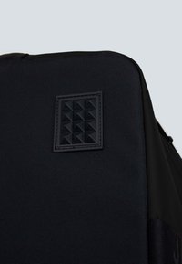 Zaino nero con un tessuto texture e un pannello nero opaco e liscio. Marcata toppa quadrata con borchie a piramide su un lato.
