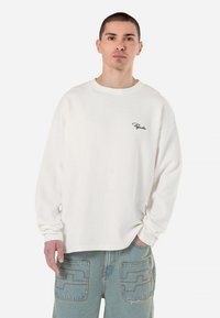 Hvid waffle-strikket langærmet skjorte med rund hals og logo broderi på venstre bryst. Sammen med lyseblå jeans med baglommer.