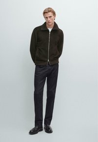 Giacca marrone con zip e maniche a costine, abbinata a pantaloni scuri e scarpe nere. Camicia bianca semplice sotto. Texture liscia in tutto.