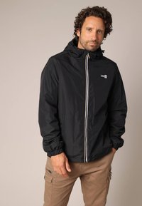 Man met krullend haar in een zwarte zip-up hoodie en beige cargobroek, staand met één hand in de broekzak tegen een effen achtergrond.