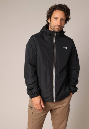 Man with curly hair wearing a black zip-up hooded jacket and beige cargo pants, standing with one hand in pocket against a plain background.