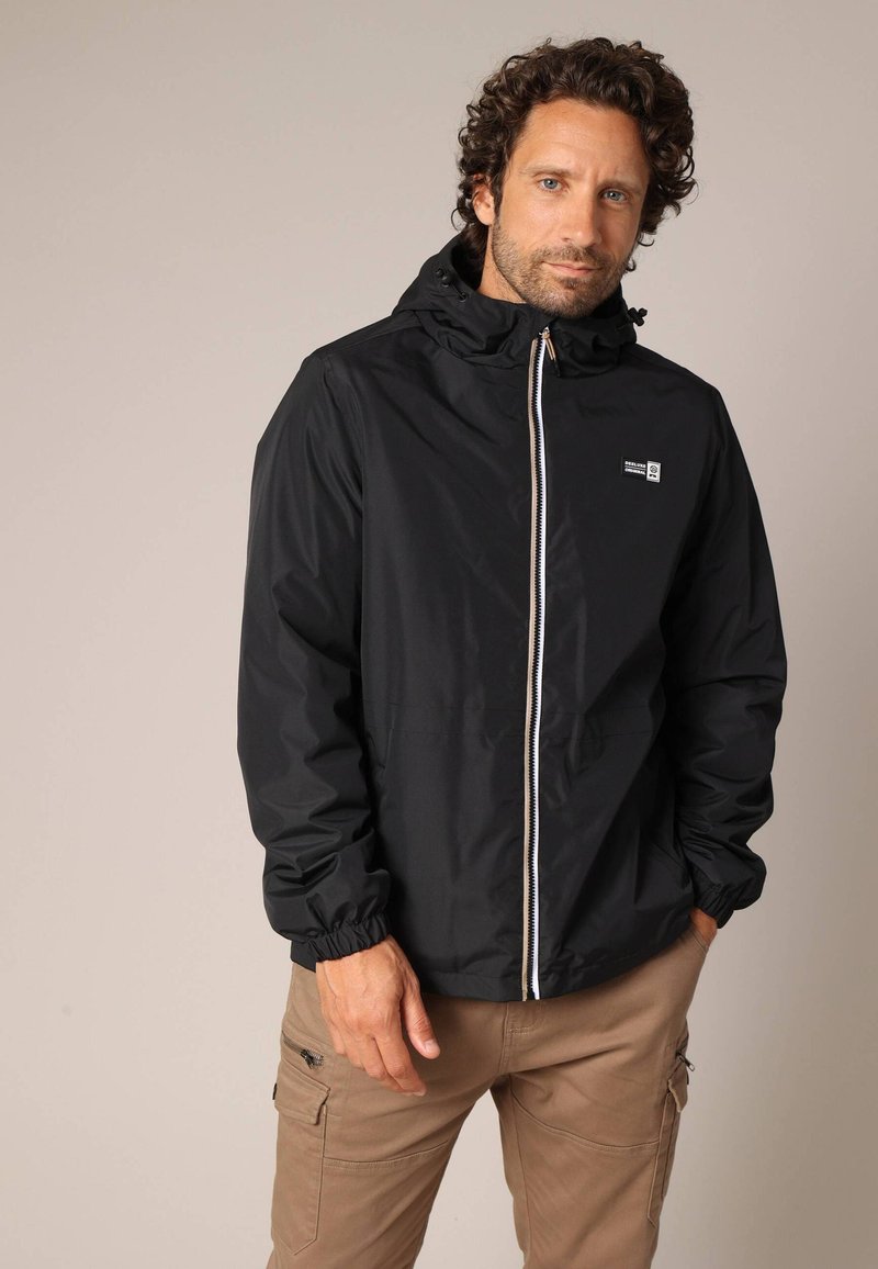 Man met krullend haar in een zwarte zip-up hoodie en beige cargobroek, staand met één hand in de broekzak tegen een effen achtergrond.