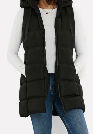 Vrouw draagt een zwarte doorgestikte mouwloze capuchonvest over een witte top met lange mouwen en blauwe spijkerbroek, handen in de zakken van de vest.