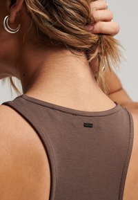 Top sin mangas color marrón con un escote redondo y una pequeña etiqueta de logo negro en la parte posterior. La tela parece suave con una textura lisa.