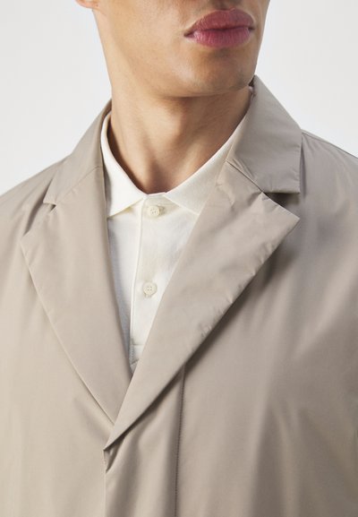Beige lightweight jacket with a notched collar, double-breasted design, and smooth texture, layered over a white collared shirt.