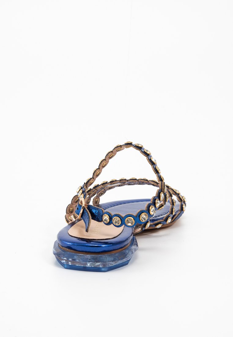 Sandali blu piatti con accenti metallici oro, caratterizzati da una cintura decorata di pietre preziose e un tacco ottagonale trasparente. Il design è minimalista con una texture liscia.