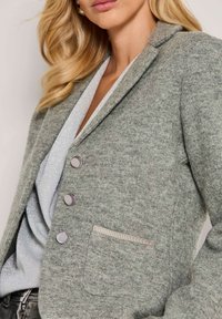 Blonde vrouw met een grijze gewatteerde blazer met drie ronde knopen over een zilver glinsterend top.
