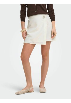 Žena nosí krémovú zavinovaciu sukňu s veľkým gombíkom, hnedé tričko s dlhým rukávom, béžové pletené slingback baleríny a krémový náramok.